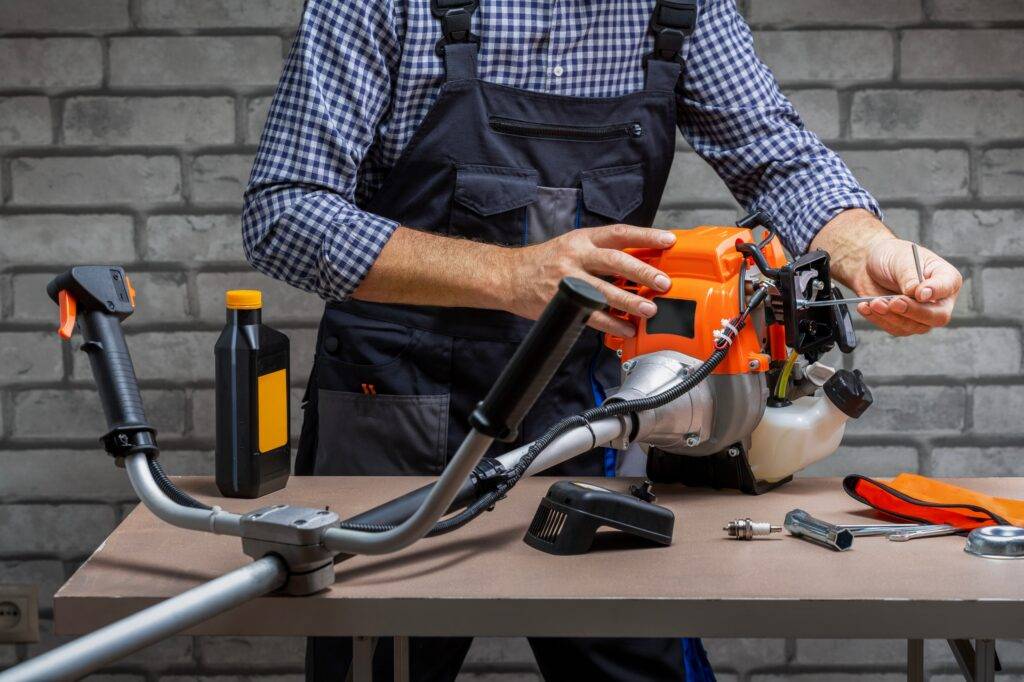 Person repairing a small engine on a workbench, holding tools and surrounded by oil and parts, illustrating lawn mower troubleshooting and maintenance for Powerline Equipment.