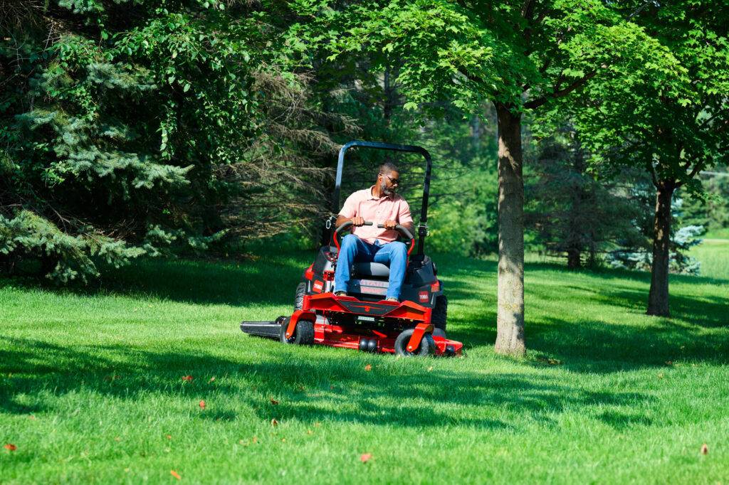 Man operating a Toro riding mower on a lush green lawn surrounded by trees, demonstrating features relevant to residential mowing.