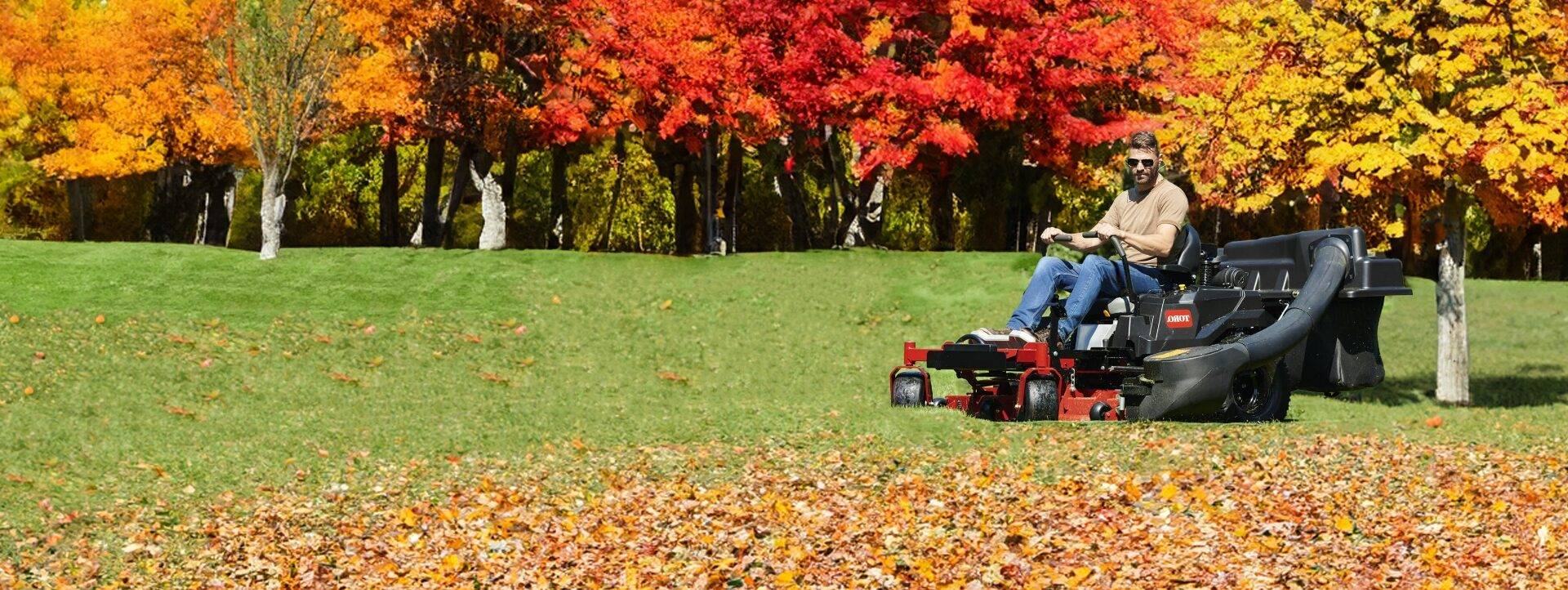 Person riding a riding mower on a grassy field covered with autumn leaves, surrounded by colorful fall foliage, illustrating lawn care for seasonal maintenance.